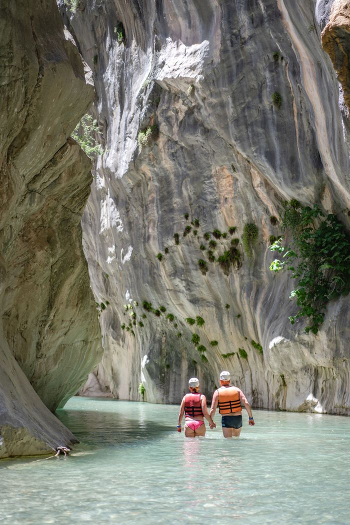 Two adults explore a scenic canyon, wearing life jackets in clear waters, surrounded by rocky cliffs.