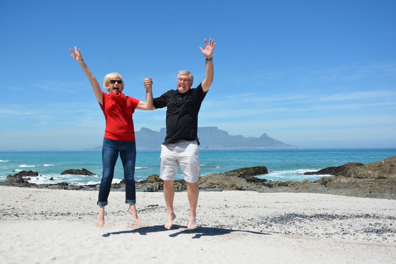 mobile-02 Elderly couple jumping with joy on a beach in Cape Town with Table Mountain in the background.