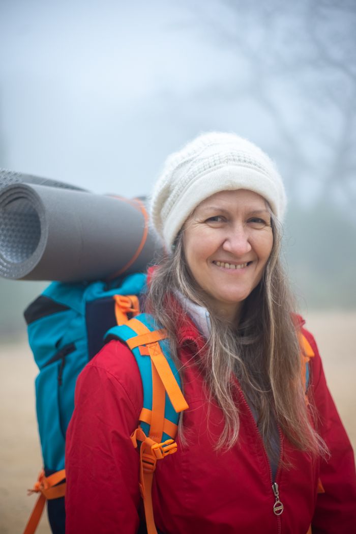 brand-02 Elderly woman smiling with backpack and camping gear in a misty outdoor environment.