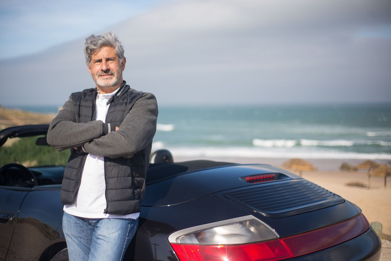 creative-02 An elderly man stands confidently by his car on a scenic beach in Portugal.